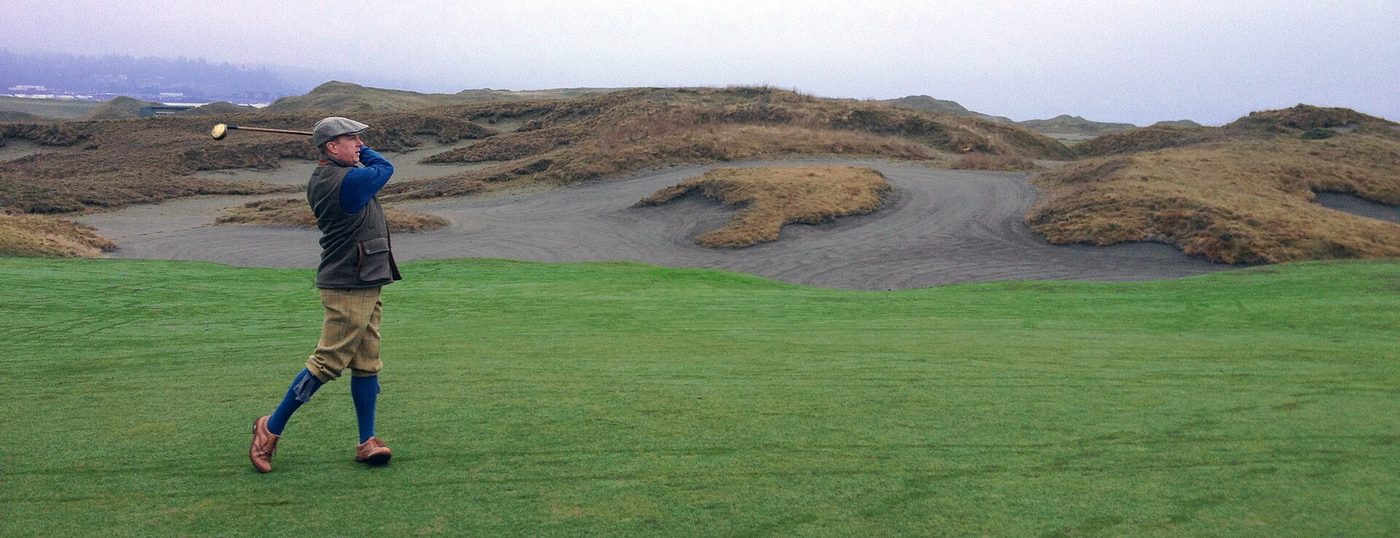 A golfer in plus-fours hitting a hickory-shafted club on the 14th hole at Chambers Bay among fescue-covered dunes