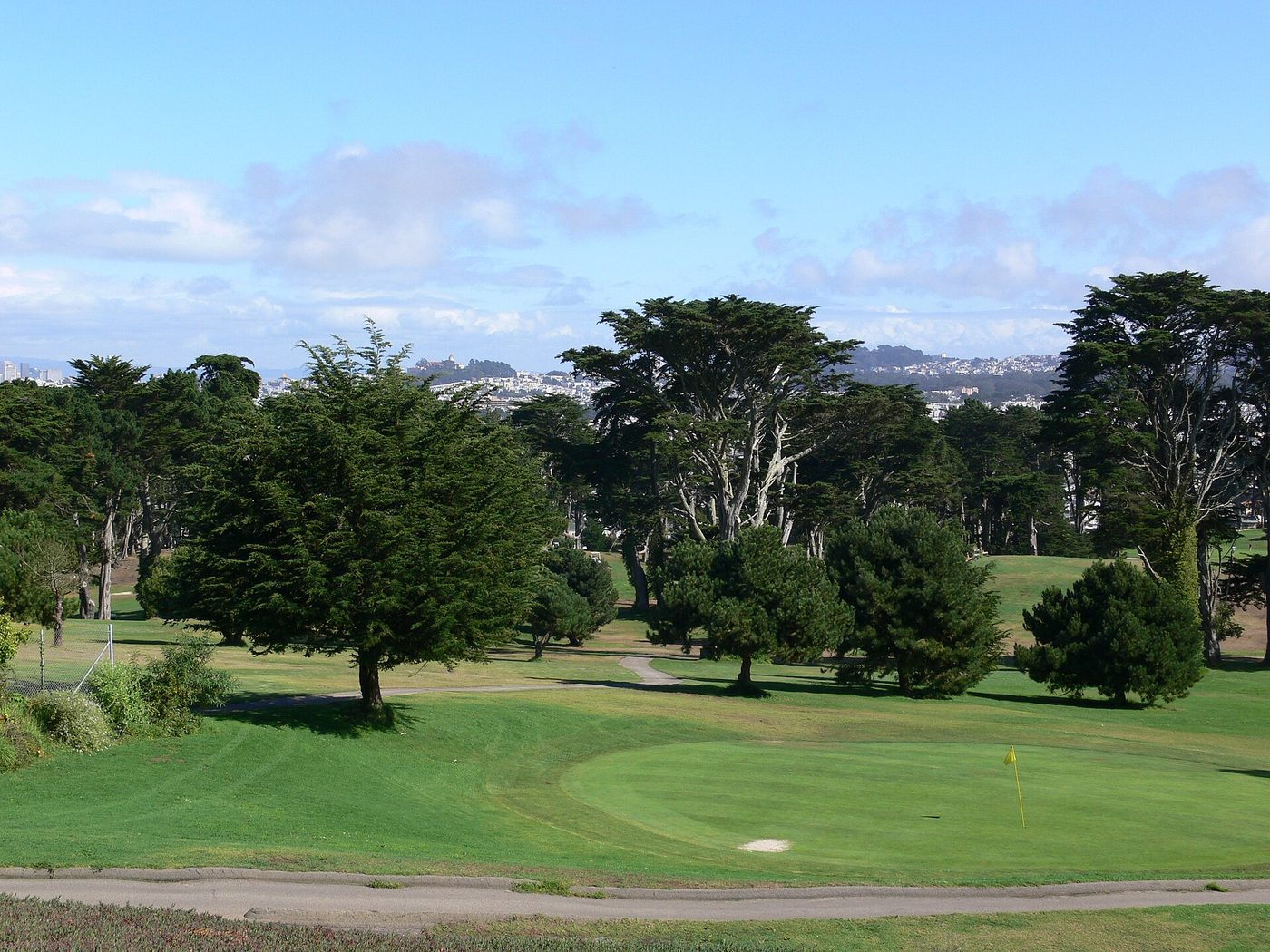 A green framed by cypress trees at Lincoln Park Golf Course with the San Francisco skyline behind