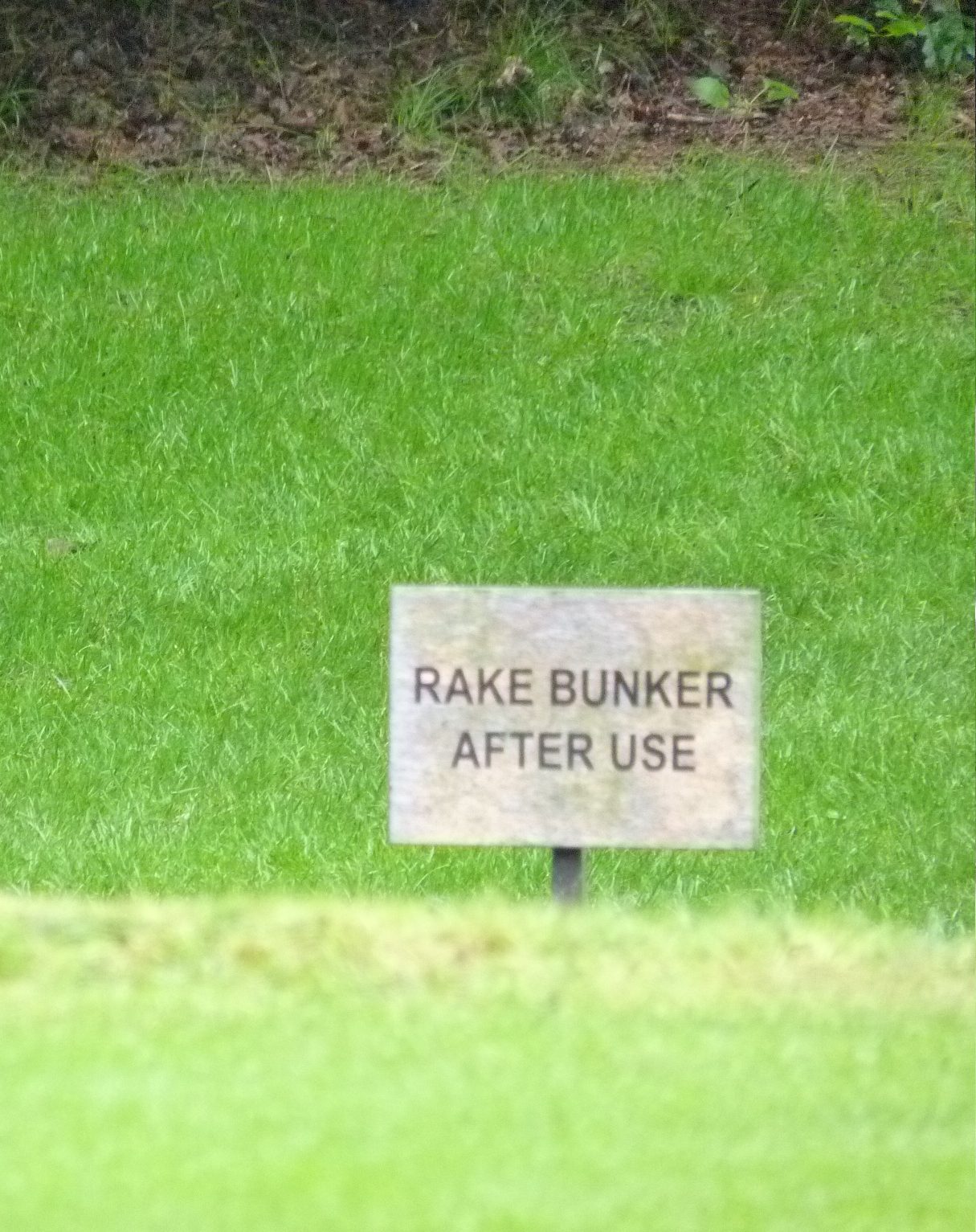 A stone sign reading Rake bunker after use beside a bunker on a golf course