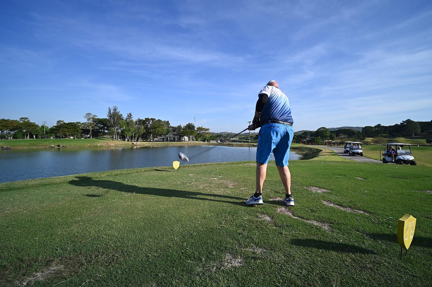 A golfer hits a tee shot between yellow tee markers over a lake, with carts on the path behind