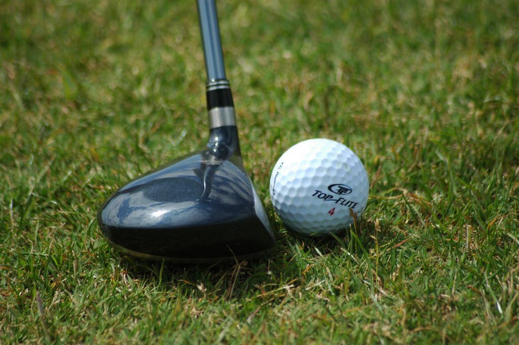 A golf ball sitting on short grass beside the head of a hybrid club, the kind of closely mown lie preferred lies apply to