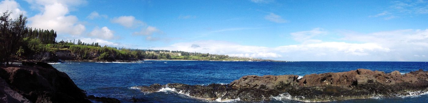 A panorama of the rocky lava shoreline and blue water at Kapalua on Maui, with the wooded coast curving away