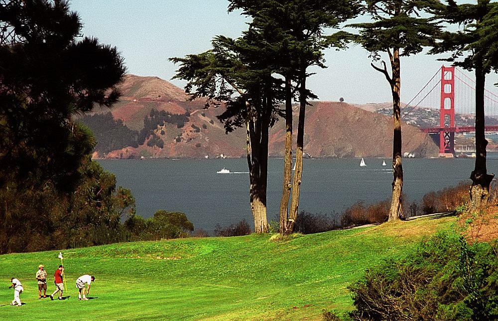 Golfers on a fairway at Lincoln Park Golf Course in San Francisco, with cypress trees and the Golden Gate Bridge behind
