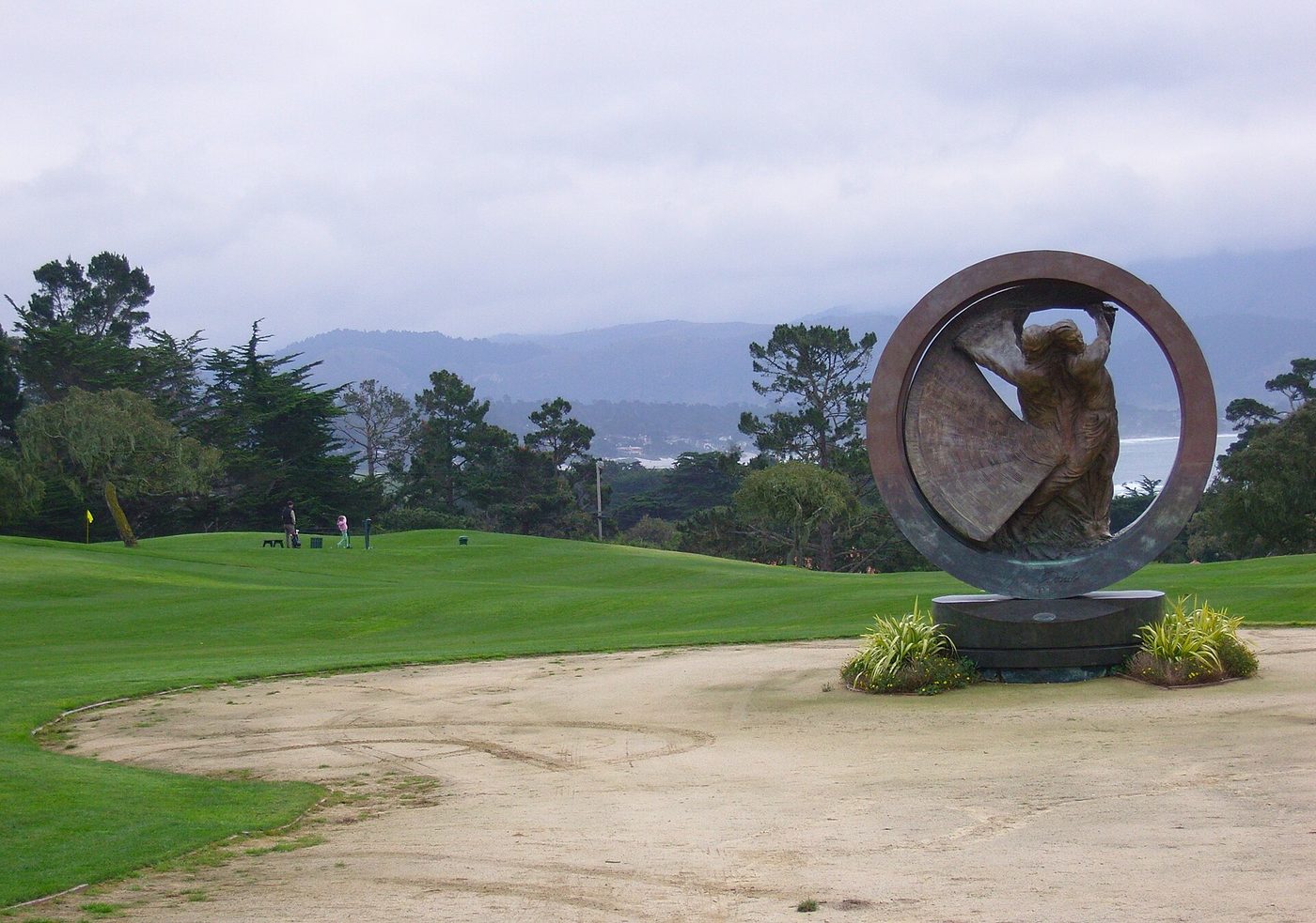 A bronze circular sculpture of a golfer beside the short course at Pebble Beach, with fairways and the coast behind